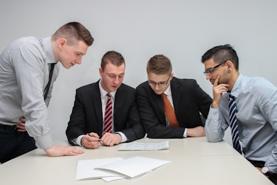 A group of four men in business attire are gathered around a table, examining documents laid out in front of them. They appear to be engaged in a discussion or meeting, with focused expressions and attentive body language.