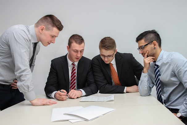 A group of four men in business attire are gathered around a table, examining documents laid out in front of them. They appear to be engaged in a discussion or meeting, with focused expressions and attentive body language.