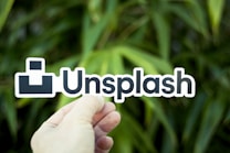 A hand holding a sticker or sign displaying the 'Unsplash' logo against a background of green foliage. The background consists of blurry, lush green plants, suggesting an outdoor setting. The logo is in bold black text with a simple geometric design in black and white.