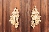 Two ornate gold door knockers with floral patterns are mounted on a rich, dark wooden door. The intricate design reflects light, highlighting their elegance and craftsmanship.
