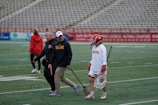 A group of people are on a sports field. One person is dressed in lacrosse gear, including a helmet and a stick, while others are wearing casual sportswear. The bleachers in the background are empty, and there are advertisements along the fences. The atmosphere suggests a practice or a pre-game warm-up.