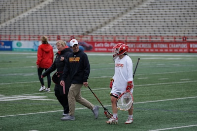 A group of people are on a sports field. One person is dressed in lacrosse gear, including a helmet and a stick, while others are wearing casual sportswear. The bleachers in the background are empty, and there are advertisements along the fences. The atmosphere suggests a practice or a pre-game warm-up.