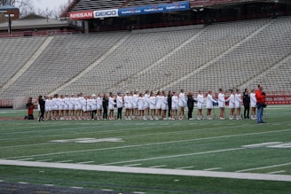 A group of athletes dressed in white and red uniforms stand in a line on a sports field, likely preparing for a game. They are being photographed by several people, while the empty bleachers in the background suggest an upcoming event.