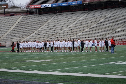 A group of athletes dressed in white and red uniforms stand in a line on a sports field, likely preparing for a game. They are being photographed by several people, while the empty bleachers in the background suggest an upcoming event.