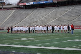 A group of athletes dressed in white and red uniforms stand in a line on a sports field, likely preparing for a game. They are being photographed by several people, while the empty bleachers in the background suggest an upcoming event.