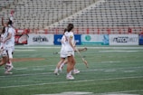A group of female lacrosse players in full gear walking across a field. They are wearing matching white uniforms with red accents and holding lacrosse sticks. The stadium seats in the background are mostly empty, but there are advertisement boards visible along the fence.
