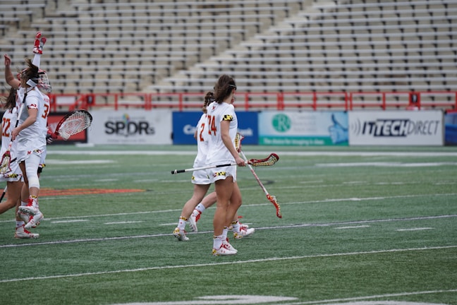 A group of female lacrosse players in full gear walking across a field. They are wearing matching white uniforms with red accents and holding lacrosse sticks. The stadium seats in the background are mostly empty, but there are advertisement boards visible along the fence.