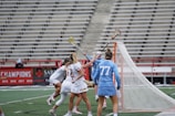 A group of female lacrosse players is engaged in an intense game. They are near the goal post with one player in a light blue uniform defending against opponents in white uniforms with red and black accents. A player is raising her lacrosse stick, seemingly trying to make a play.