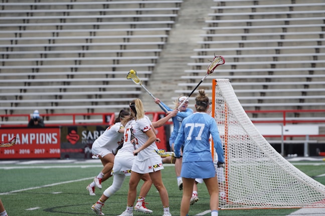 A group of female lacrosse players is engaged in an intense game. They are near the goal post with one player in a light blue uniform defending against opponents in white uniforms with red and black accents. A player is raising her lacrosse stick, seemingly trying to make a play.