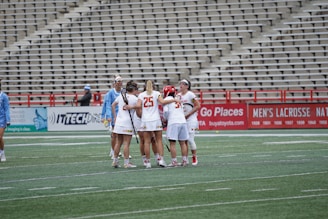 A group of female lacrosse players wearing uniforms gather together on a sports field, surrounded by an empty stadium. The players appear to be huddling, possibly discussing or strategizing during a match. Their uniforms are mostly white with red accents. Another group of players in light blue uniforms can be seen nearby.