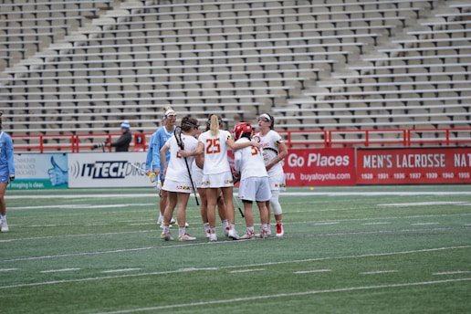 A group of female lacrosse players wearing uniforms gather together on a sports field, surrounded by an empty stadium. The players appear to be huddling, possibly discussing or strategizing during a match. Their uniforms are mostly white with red accents. Another group of players in light blue uniforms can be seen nearby.