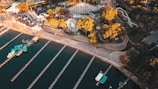 An aerial view of an amusement park located near a body of water. The amusement park features a Ferris wheel, roller coaster tracks, and various other rides. The park is surrounded by lush trees that have an autumnal golden hue. Along the water, there are several docks and boats, with the piers extending out into the water. Roads and pathways can be seen encircling the park area.
