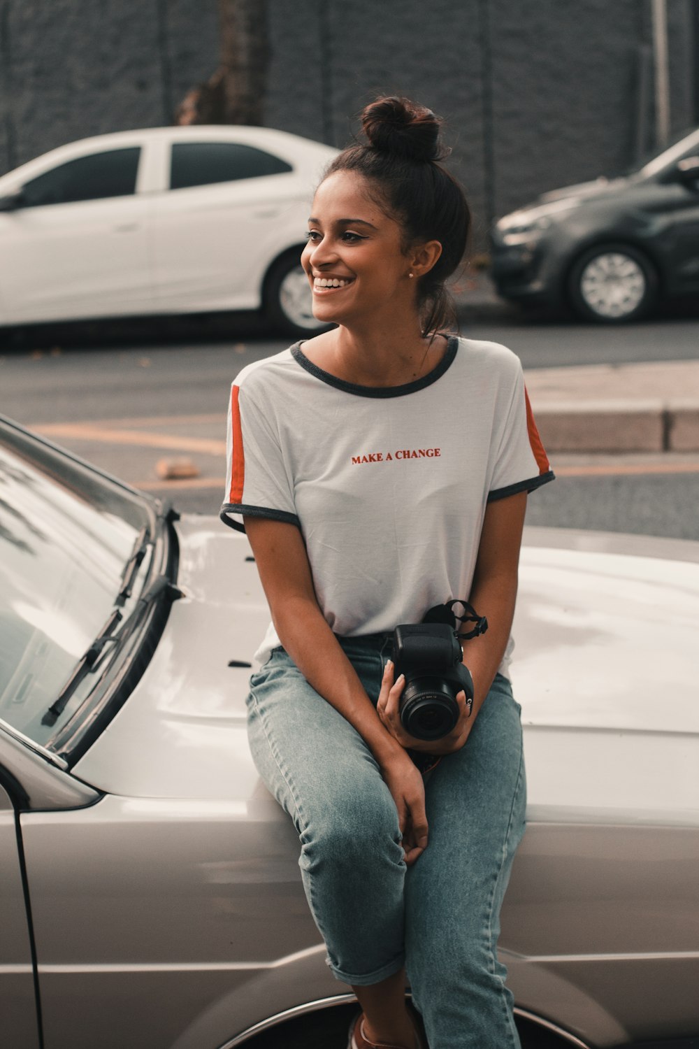 smiling woman sits on car hood holding camera