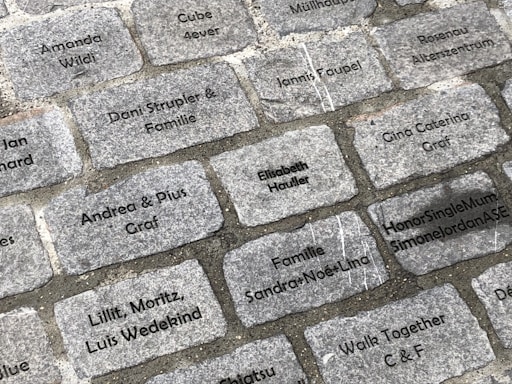 Engraved cobblestone bricks are laid out in a rectangular pattern. Each brick contains inscriptions of different names or phrases such as 'Amanda Wildi', 'Andrea & Pius Graf', 'HonorSingleMumSimoneJordanASE', and 'Familie Sandra-Noe-Lina'. The inscriptions are in black text on gray stones.