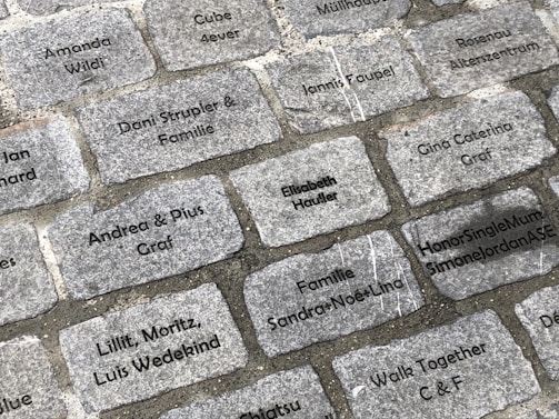 Engraved cobblestone bricks are laid out in a rectangular pattern. Each brick contains inscriptions of different names or phrases such as 'Amanda Wildi', 'Andrea & Pius Graf', 'HonorSingleMumSimoneJordanASE', and 'Familie Sandra-Noe-Lina'. The inscriptions are in black text on gray stones.