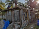 The image depicts a construction site with workers building a small structure made of concrete blocks. There are wooden scaffolding and support beams in place, and the site is surrounded by palm trees, conveying a tropical setting. Construction materials such as blue tarpaulin and concrete blocks are scattered around the area.