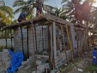 The image depicts a construction site with workers building a small structure made of concrete blocks. There are wooden scaffolding and support beams in place, and the site is surrounded by palm trees, conveying a tropical setting. Construction materials such as blue tarpaulin and concrete blocks are scattered around the area.