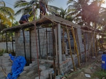 The image depicts a construction site with workers building a small structure made of concrete blocks. There are wooden scaffolding and support beams in place, and the site is surrounded by palm trees, conveying a tropical setting. Construction materials such as blue tarpaulin and concrete blocks are scattered around the area.