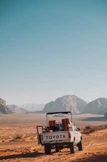 A white Toyota pickup truck is parked on a vast desert landscape with reddish sand. Mountains are visible in the background under a clear blue sky. The truck bed contains various items, including red and black cushions or bags. Tracks in the sand suggest movement.