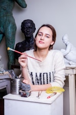 A woman is seated at an art studio, holding a paintbrush. She is surrounded by sculptures and sculpting tools. She wears a sweater with the word 'AMOUR' printed on it. The workspace is filled with artistic materials, and several statues are visible in the background.
