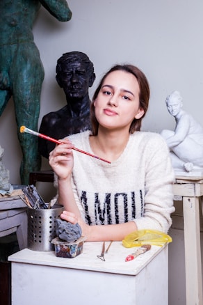 A woman is seated at an art studio, holding a paintbrush. She is surrounded by sculptures and sculpting tools. She wears a sweater with the word 'AMOUR' printed on it. The workspace is filled with artistic materials, and several statues are visible in the background.