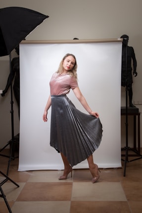 A woman in a pink top and metallic silver pleated skirt poses gracefully in front of a white backdrop. She is standing on a beige and brown tiled floor. There is a large black studio light on a stand to the left side and another stand visible to the right. In the background, two dark statues add a dramatic contrast to the scene.
