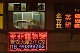 A storefront window at night displays several illuminated signs in a foreign script, likely an Asian language. Inside the window display, a cat sleeps on a platform within a cage, while another cat stands nearby. The scene is dimly lit except for the bright signage, which includes various advertisements and contact information.