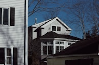 A residential scene depicting two houses with white siding and dark shutters. The main house features large windows on the second floor, and trees without leaves frame the setting. The sky is clear and blue.