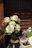 A garden scene with ornamental cabbage plants and other flowers in pots. The main plant has large cream and green leaves, while the potted flowers beside it are pink and white. Wooden slats in the background provide a rustic ambiance.