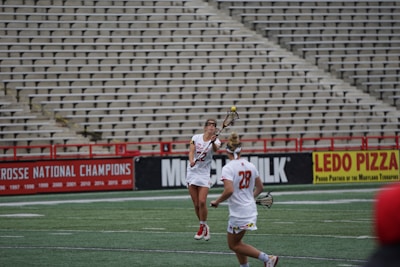 Two female athletes in white uniforms with red accents are playing lacrosse on a field. One is in motion catching a ball with a stick, while an empty stadium with rows of seats is in the background. Advertisements for Lacrosse National Champions and a pizza company are displayed on the stands.