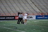 A player in a white sports uniform with the number 37 is standing on a sports field, accompanied by two referees in black and white striped shirts. The background shows empty stadium seating and advertising banners along the sidelines.