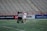 A player in a white sports uniform with the number 37 is standing on a sports field, accompanied by two referees in black and white striped shirts. The background shows empty stadium seating and advertising banners along the sidelines.