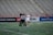 A player in a white sports uniform with the number 37 is standing on a sports field, accompanied by two referees in black and white striped shirts. The background shows empty stadium seating and advertising banners along the sidelines.