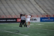 A player in a white sports uniform with the number 37 is standing on a sports field, accompanied by two referees in black and white striped shirts. The background shows empty stadium seating and advertising banners along the sidelines.