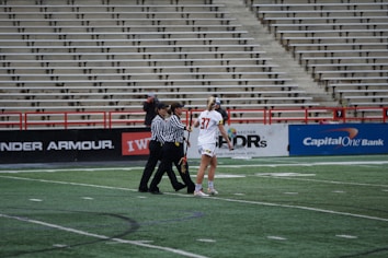 A player in a white sports uniform with the number 37 is standing on a sports field, accompanied by two referees in black and white striped shirts. The background shows empty stadium seating and advertising banners along the sidelines.