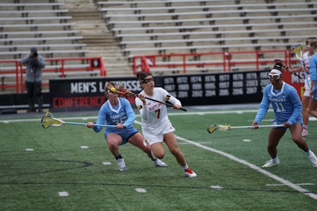 Athletes engaged in a competitive lacrosse match on a field with green turf. The players are wearing uniforms with Maryland and Carolina insignias, focusing intensely on the game. The background shows stadium seating with a few spectators.