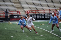 Athletes engaged in a competitive lacrosse match on a field with green turf. The players are wearing uniforms with Maryland and Carolina insignias, focusing intensely on the game. The background shows stadium seating with a few spectators.