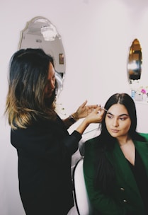 A woman is applying makeup to another woman seated in a chair. The scene takes place in a well-lit room with elegant mirrors on the wall and decorative elements. The atmosphere is professional and focused.