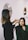 A woman is applying makeup to another woman seated in a chair. The scene takes place in a well-lit room with elegant mirrors on the wall and decorative elements. The atmosphere is professional and focused.