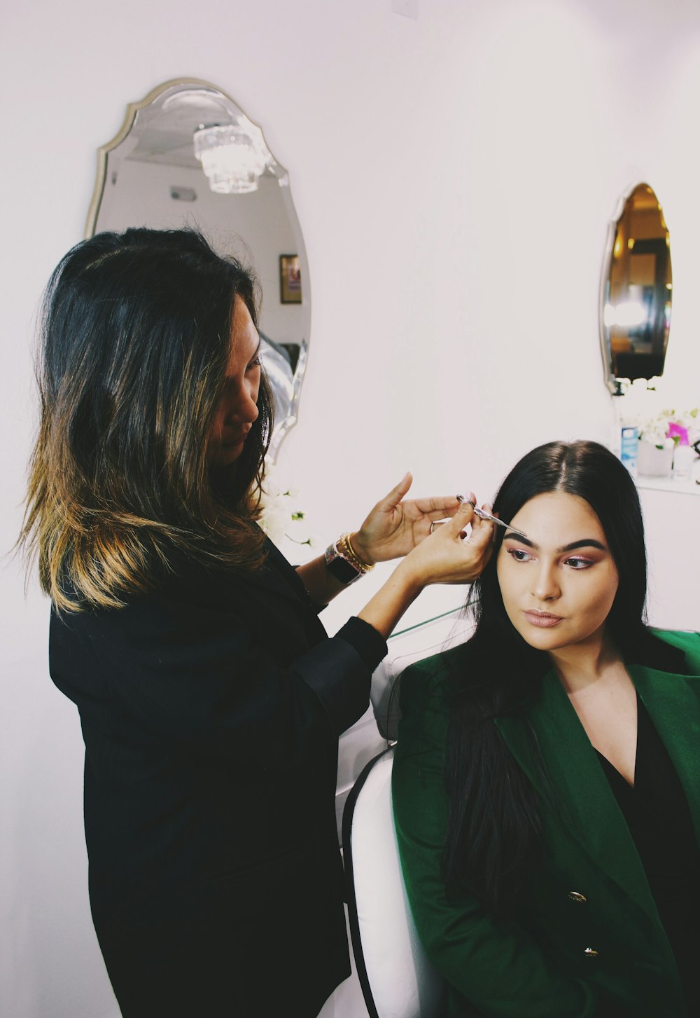 woman clipping eyebrow of woman wearing green blazer