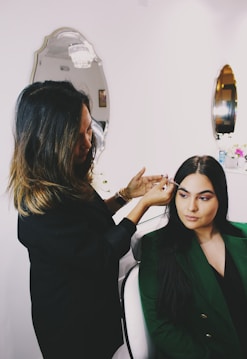 A woman is applying makeup to another woman seated in a chair. The scene takes place in a well-lit room with elegant mirrors on the wall and decorative elements. The atmosphere is professional and focused.