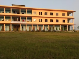 A three-story building is situated on a grassy field, featuring a pale orange facade with blue accents around doors and windows. Several plants are aligned in front of the building. A sign indicates that it is the Shilbadrar Teachers Training Institute.