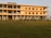 A three-story building is situated on a grassy field, featuring a pale orange facade with blue accents around doors and windows. Several plants are aligned in front of the building. A sign indicates that it is the Shilbadrar Teachers Training Institute.