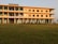 A three-story building is situated on a grassy field, featuring a pale orange facade with blue accents around doors and windows. Several plants are aligned in front of the building. A sign indicates that it is the Shilbadrar Teachers Training Institute.