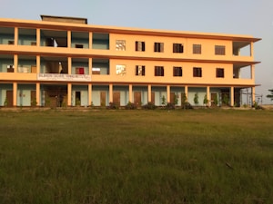 A three-story building is situated on a grassy field, featuring a pale orange facade with blue accents around doors and windows. Several plants are aligned in front of the building. A sign indicates that it is the Shilbadrar Teachers Training Institute.