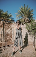 A woman stands in an outdoor setting wearing a black and white striped jumpsuit and sunglasses. She is in front of a textured, ornate metal gate and a stone wall with plants, including palm trees, in the background.