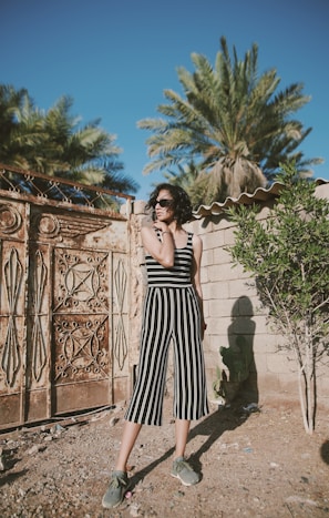 A woman stands in an outdoor setting wearing a black and white striped jumpsuit and sunglasses. She is in front of a textured, ornate metal gate and a stone wall with plants, including palm trees, in the background.