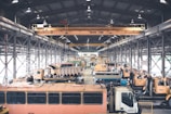 A large indoor facility filled with industrial vehicles and machinery. The space is structured with metal beams and bright overhead lighting. There are multiple heavy-duty trucks and construction equipment lined up, suggesting a maintenance or storage area. The setting appears organized with cranes and equipment systematically arranged.