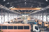 A large indoor facility filled with industrial vehicles and machinery. The space is structured with metal beams and bright overhead lighting. There are multiple heavy-duty trucks and construction equipment lined up, suggesting a maintenance or storage area. The setting appears organized with cranes and equipment systematically arranged.