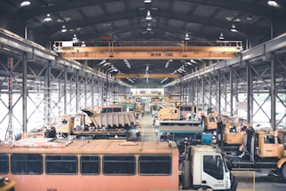 A large indoor facility filled with industrial vehicles and machinery. The space is structured with metal beams and bright overhead lighting. There are multiple heavy-duty trucks and construction equipment lined up, suggesting a maintenance or storage area. The setting appears organized with cranes and equipment systematically arranged.